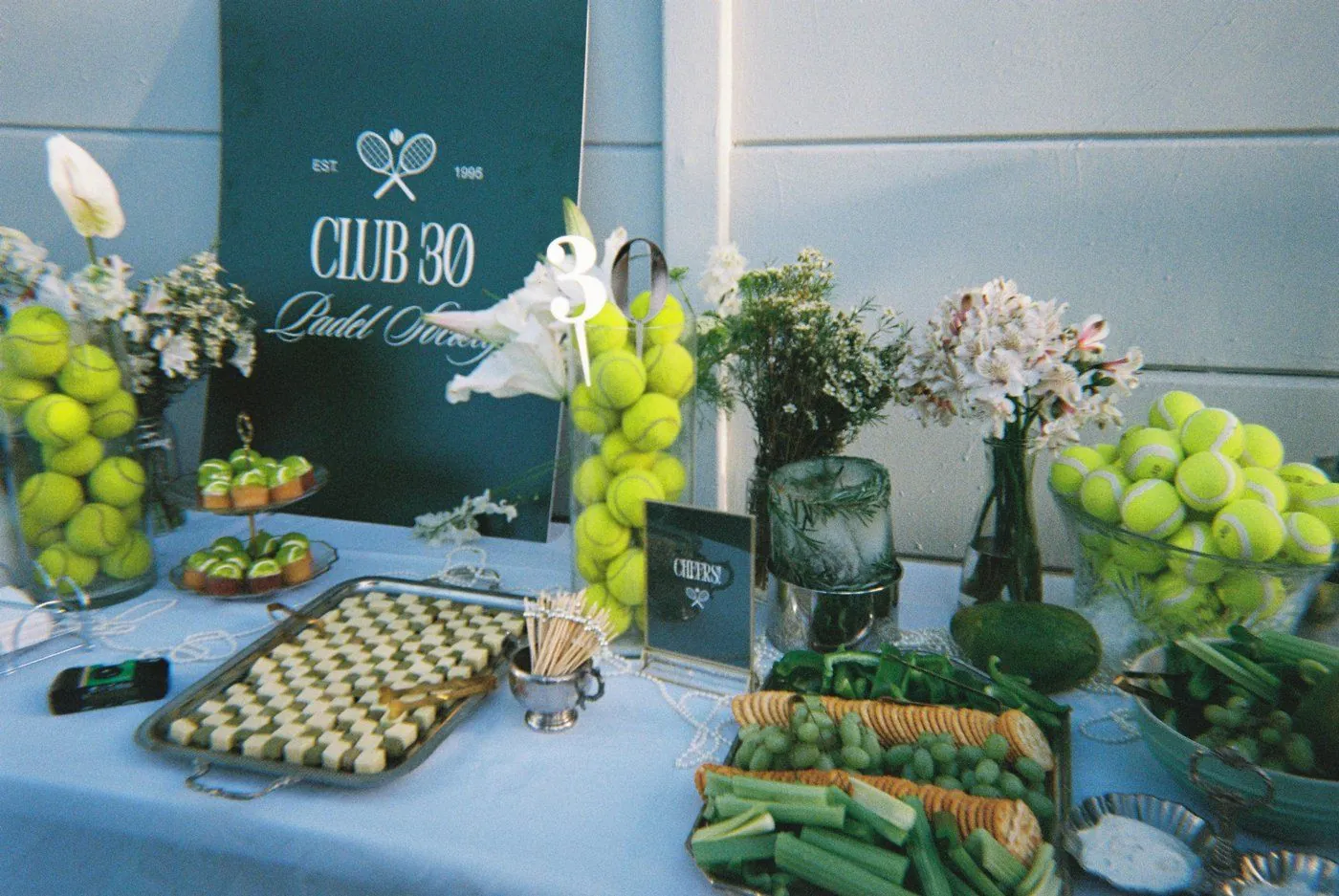 Tennis-themed Club 30 grazing table with tennis-ball arrangements, green grazing and florals