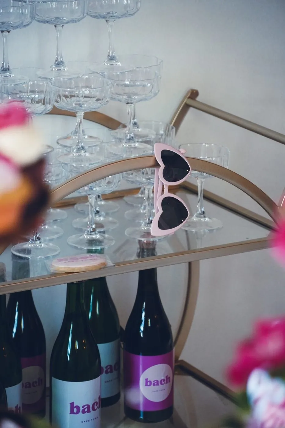 Gold bar cart stacked with coupe glasses, pink heart sunglasses and branded bach Cape Town sparkling bottles at a bachelorette