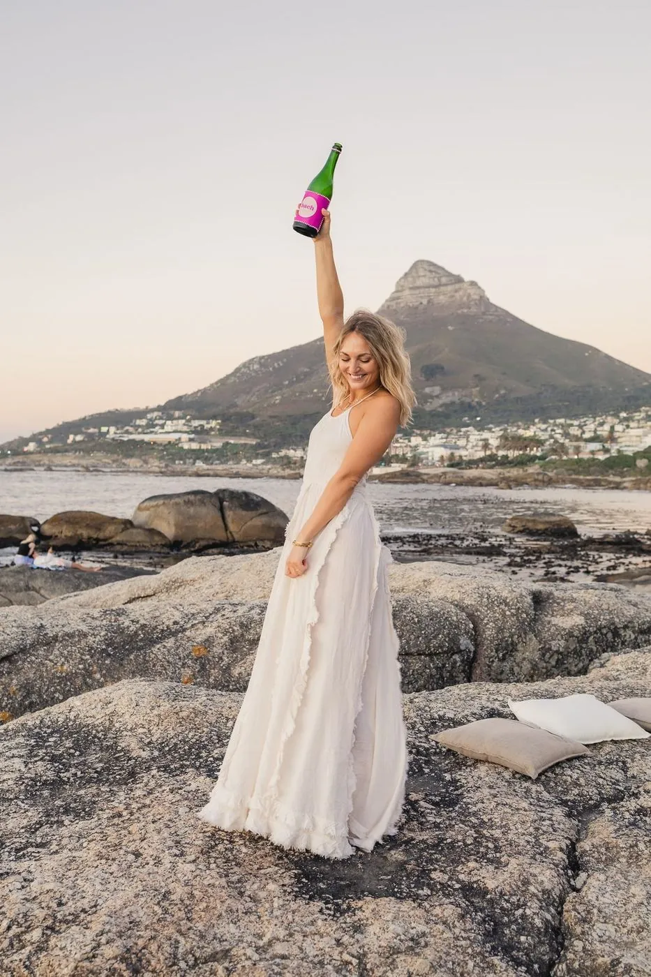 Bride in a flowing ivory dress raising a branded bach bottle on the rocks at sunset below Lion's Head, Cape Town