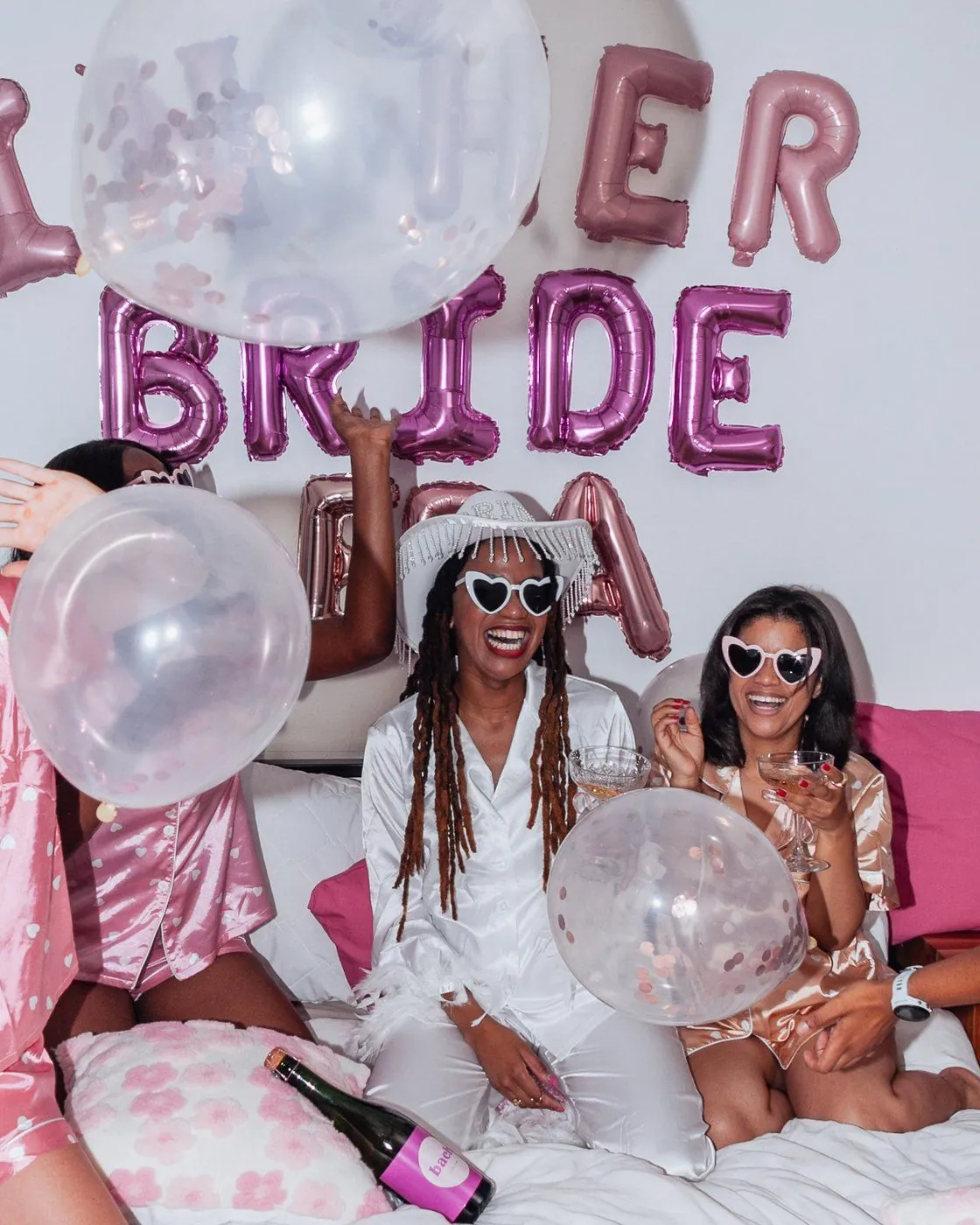 Bride and friends in satin pyjamas laughing with confetti balloons under a BRIDE balloon backdrop