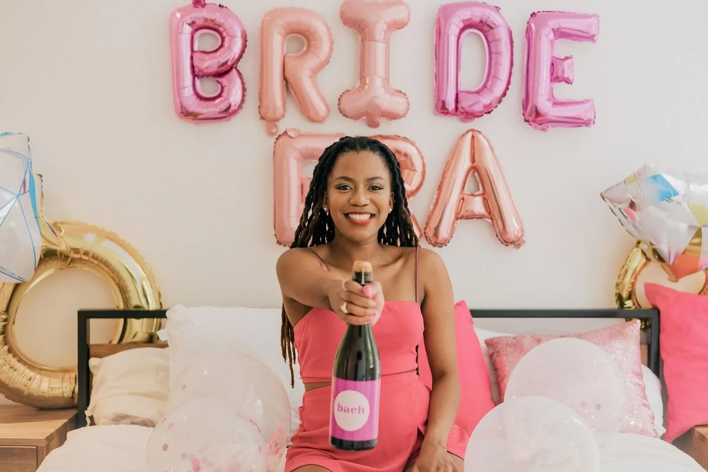 Bride-to-be in a coral dress holding a branded bach bottle beneath pink BRIDE ERA balloons at a bachelorette, Cape Town