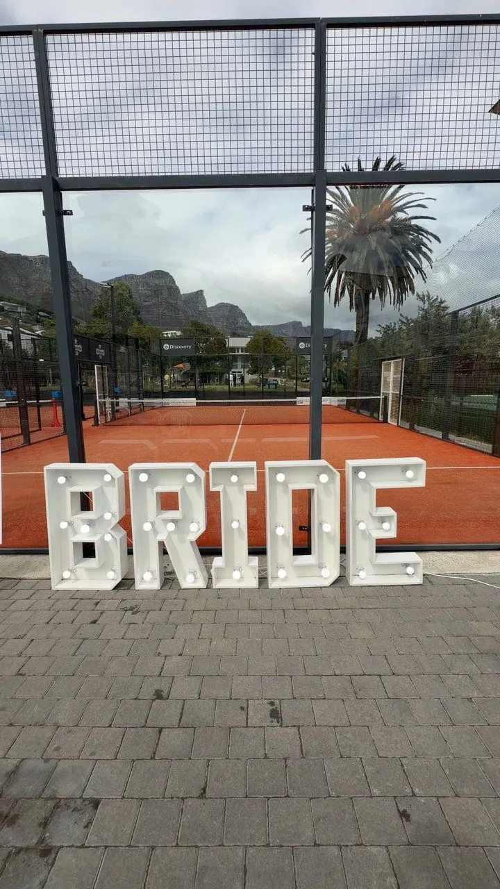 Glowing BRIDE marquee letters at a padel-court bachelorette with Table Mountain behind, Cape Town