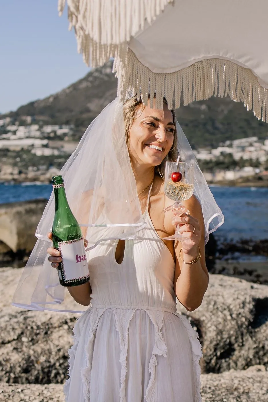 Bride in a veil holding a branded bach bottle and a cocktail under a fringed umbrella at a seaside bachelorette, Cape Town