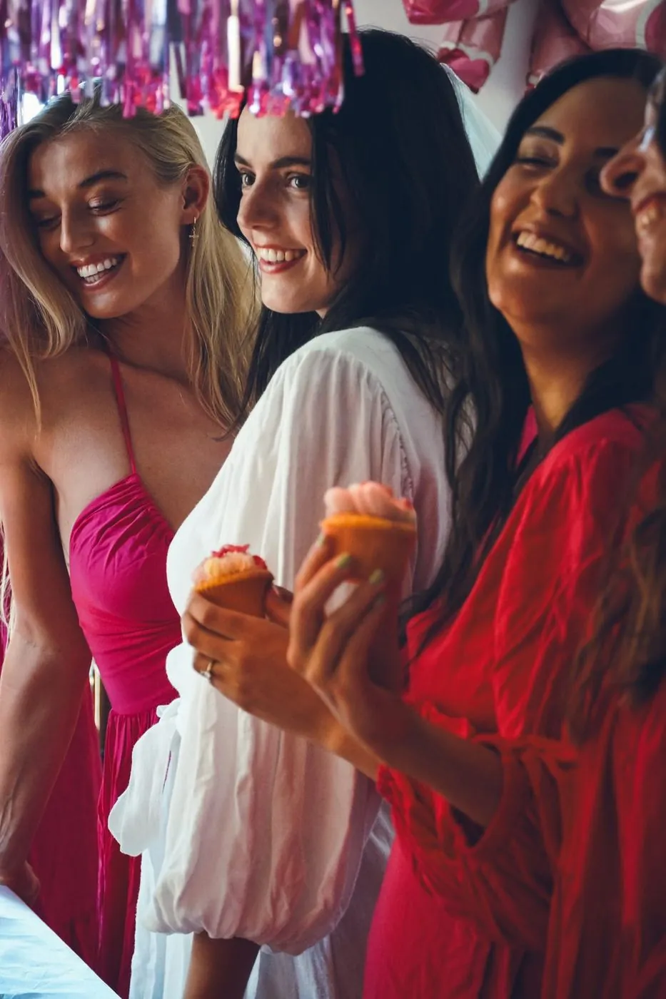Friends in pink and red laughing in a line with cupcakes under pink tinsel at a bachelorette, Cape Town