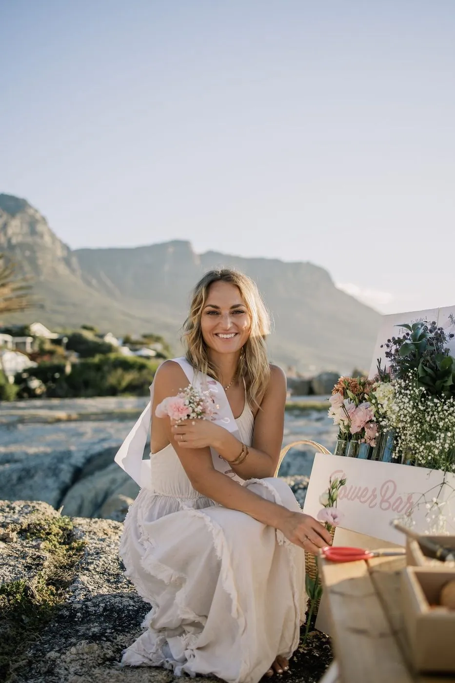 Bride-to-be in a sash seated beside a styled Flower Bar at a beachside bachelorette below Table Mountain, Cape Town