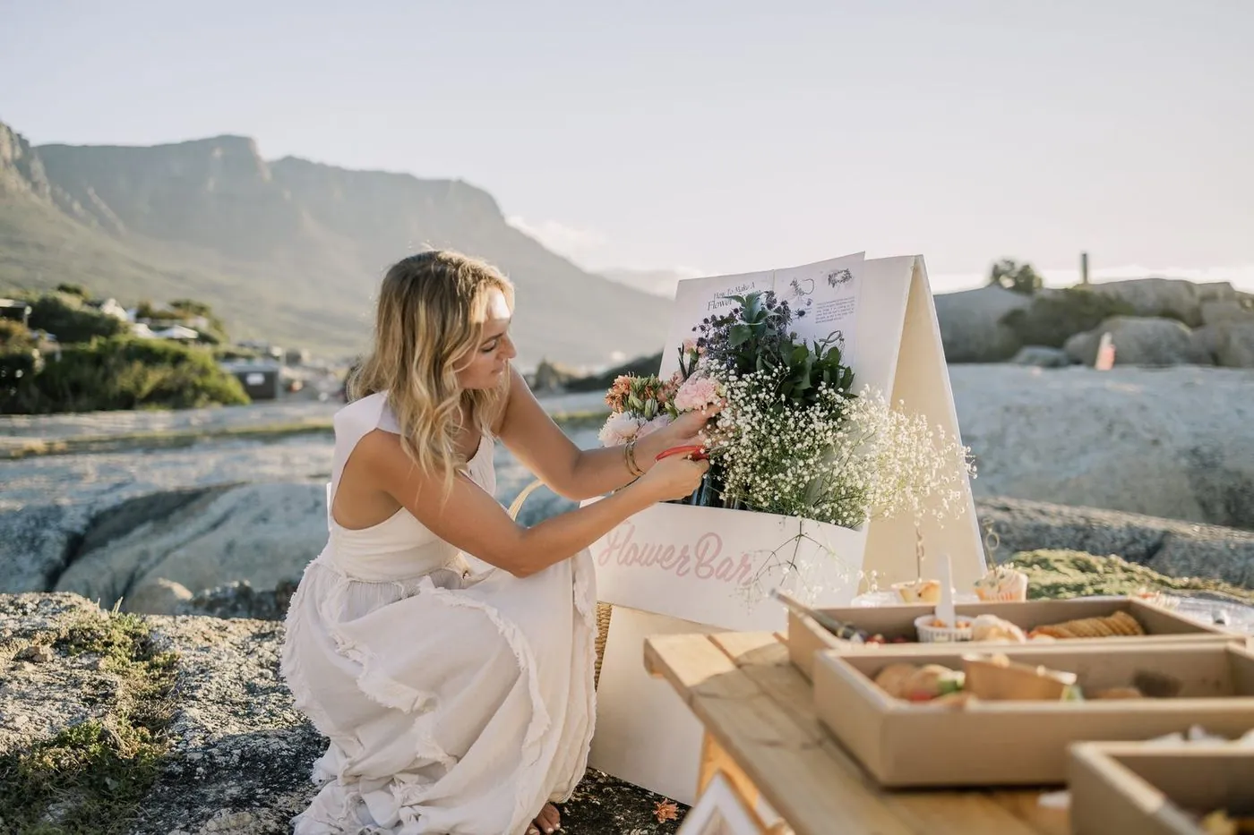 Guest arranging flowers at a seaside Flower Bar with a grazing picnic below Table Mountain, Cape Town