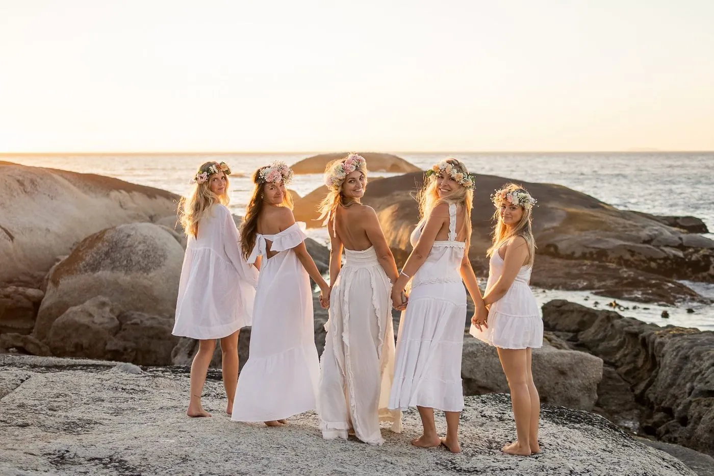 Bachelorette group in white dresses and floral crowns holding hands at golden hour by the Atlantic, Cape Town