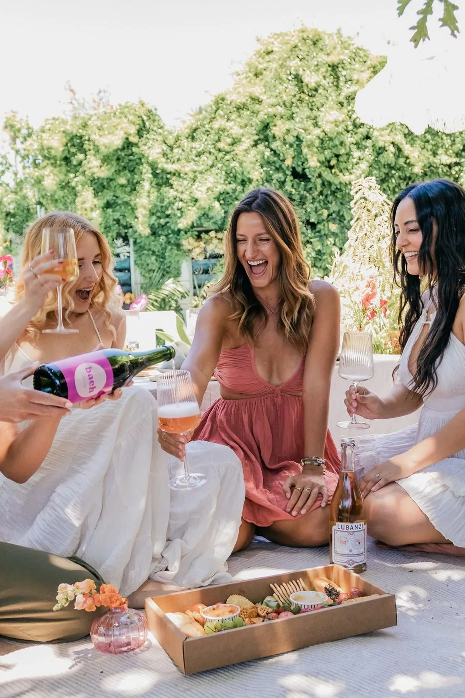 Friends laughing as branded bach sparkling is poured over a garden grazing picnic, Cape Town