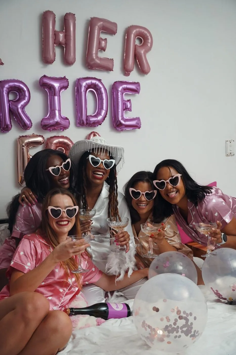 Five friends in pink satin pyjamas and heart sunglasses raising coupes under HER BRIDE ERA balloons with a branded bach bottle at a bachelorette, Cape Town