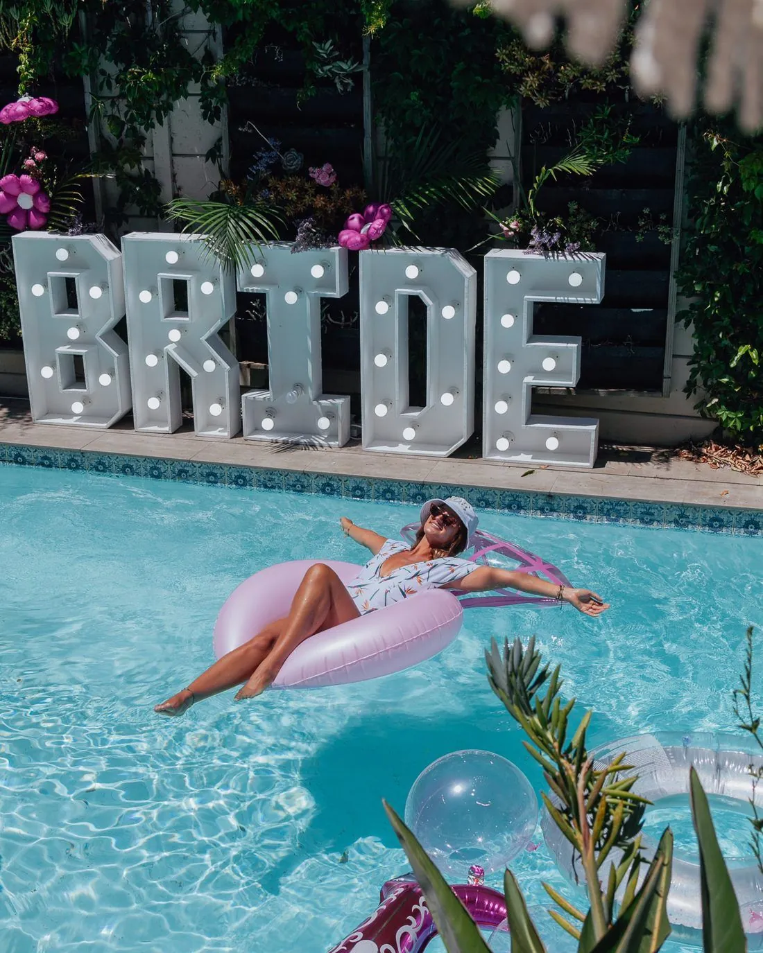 Bride relaxing on a pink float in a pool beside glowing BRIDE marquee letters at a villa bachelorette, Cape Town