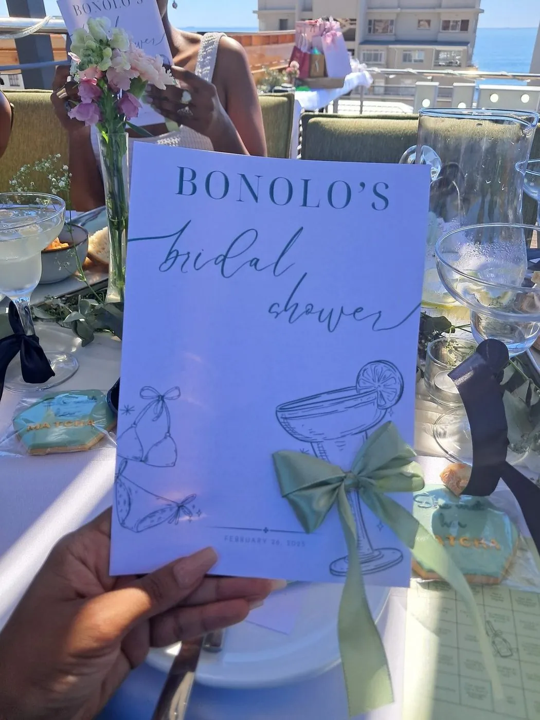 Custom bridal shower menu card with cocktail illustrations and a sage ribbon bow at a seaside table, Cape Town