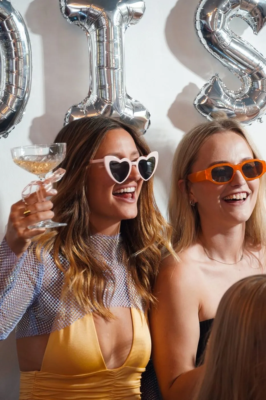 Two friends in heart and orange sunglasses raising a coupe beneath silver letter balloons at a bachelorette, Cape Town