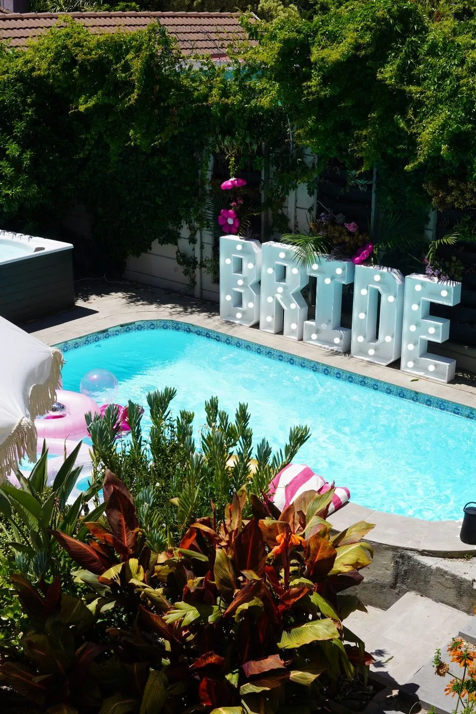 Aerial view of a villa pool with glowing BRIDE marquee letters, pink floats and a hot tub in a lush Cape Town garden
