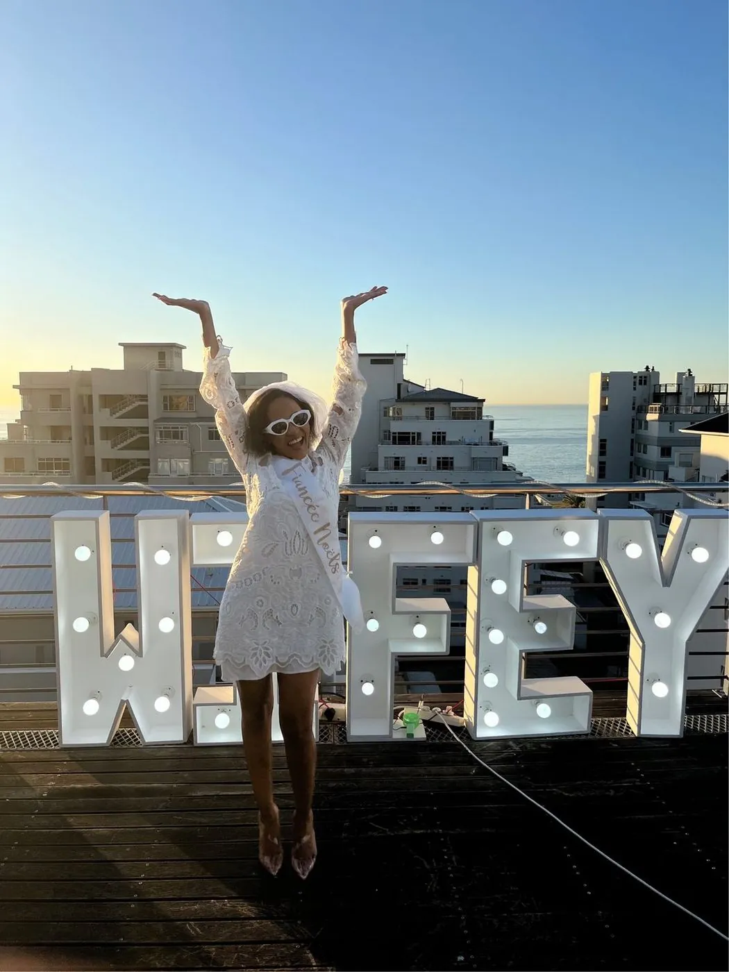 Bride-to-be in a Fiancée sash leaping for joy in front of glowing WIFEY marquee letters on a Sea Point rooftop at sunset, Cape Town